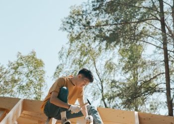 A carpenter works on a wooden roof frame outdoors under clear skies.